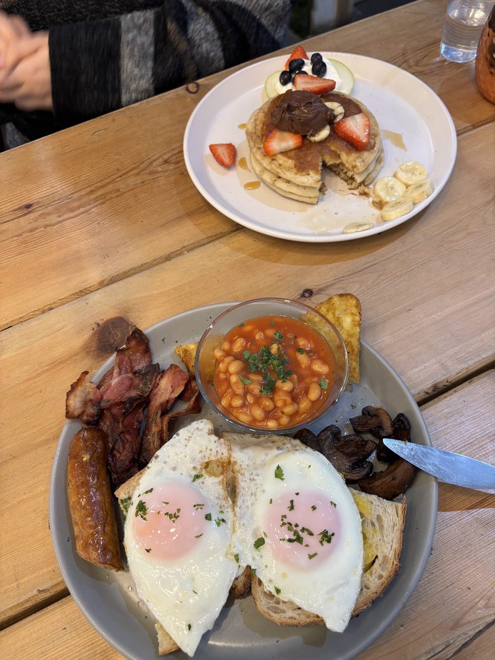 A full English breakfast with a stack of pancakes behind it on a wooden cafe table